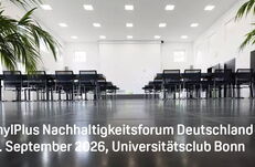 Großaufnahme eines hellen, modernen Seminarraums mit Reihen von schwarzen Stühlen und Tischen, Palmen links und rechts, mit projizierter weißer Textfolie 'VinylPlus Nachhaltigkeitsforum Deutschland 22. September 2026, Universitätsclub Bonn'